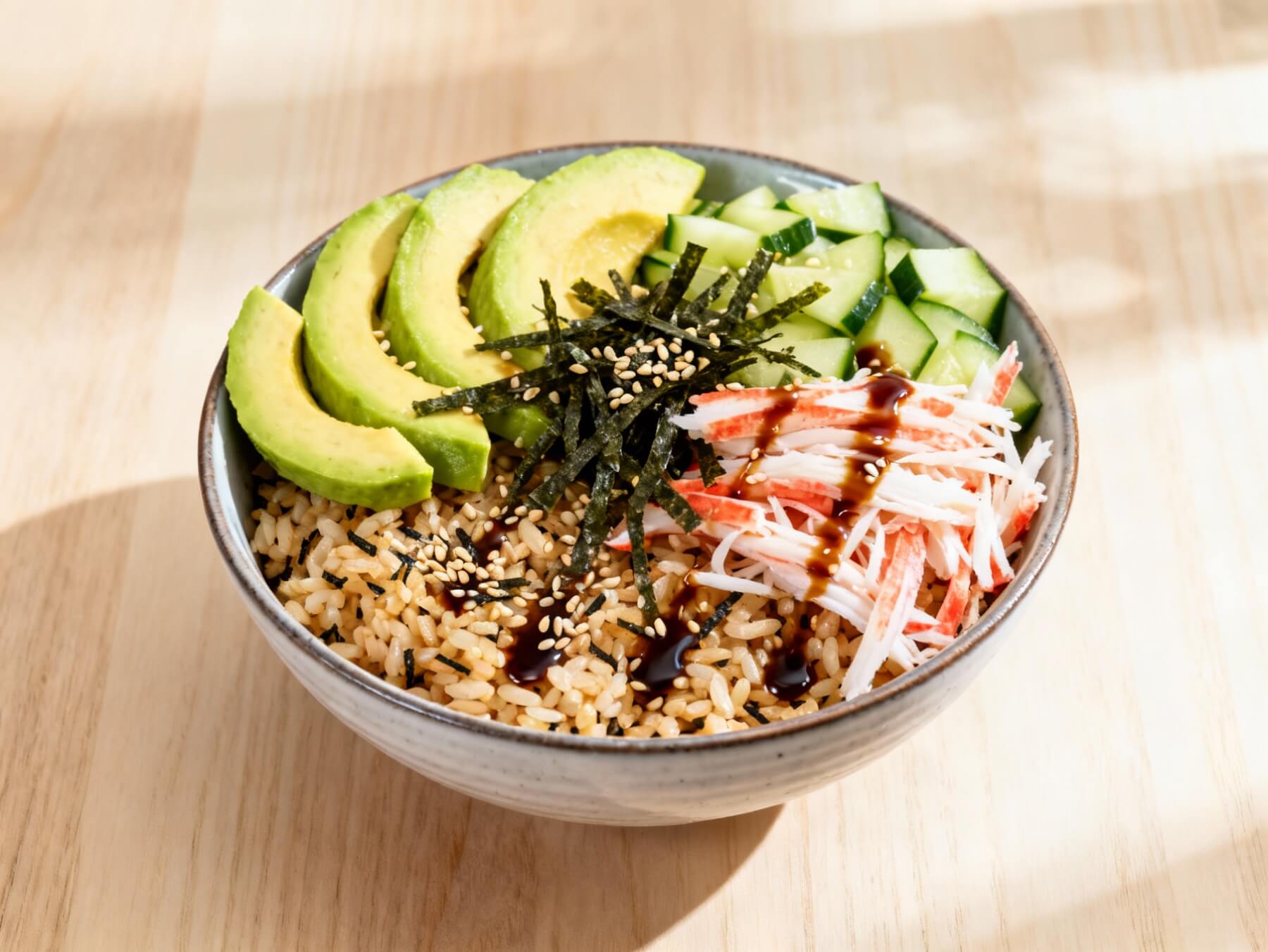 Sushi bowl with brown rice, avocado, cucumber, crab, seaweed, and sesame seeds drizzled with soy glaze.