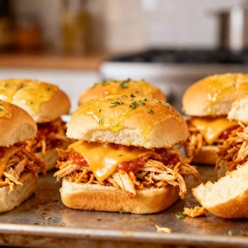 Juicy buffalo chicken sliders with shredded chicken, melted cheese, and Hawaiian rolls brushed with buttery sauce, arranged on a baking tray.