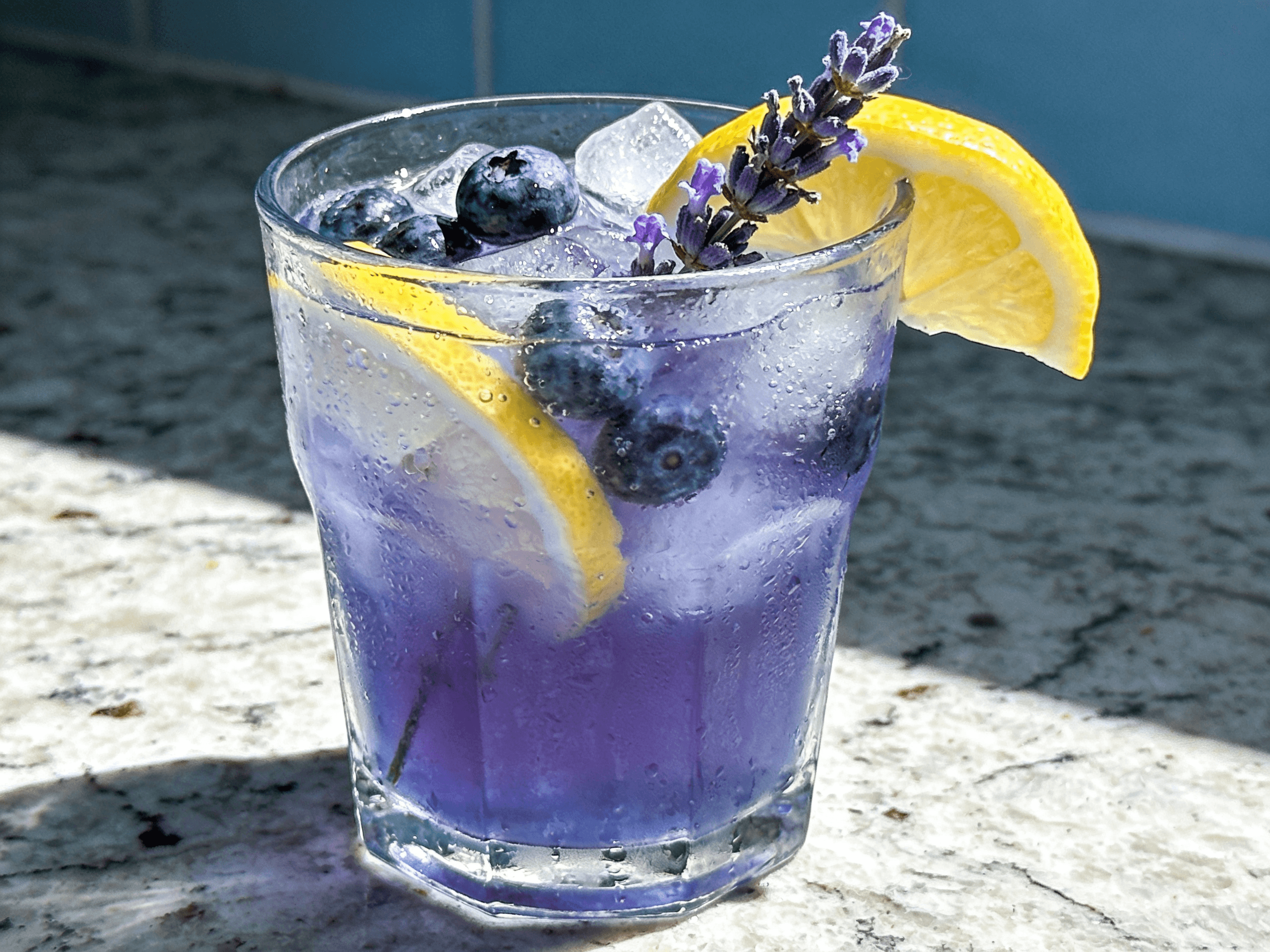 Close-up front view of Blueberry Lavender Lemonade with fresh blueberries, lemon slices, lavender sprigs, crushed ice, and chilled floral summer drink styling.