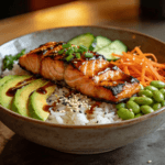 Front view of teriyaki salmon rice bowl with avocado, edamame, carrots, cucumber, and sesame seeds over jasmine rice.