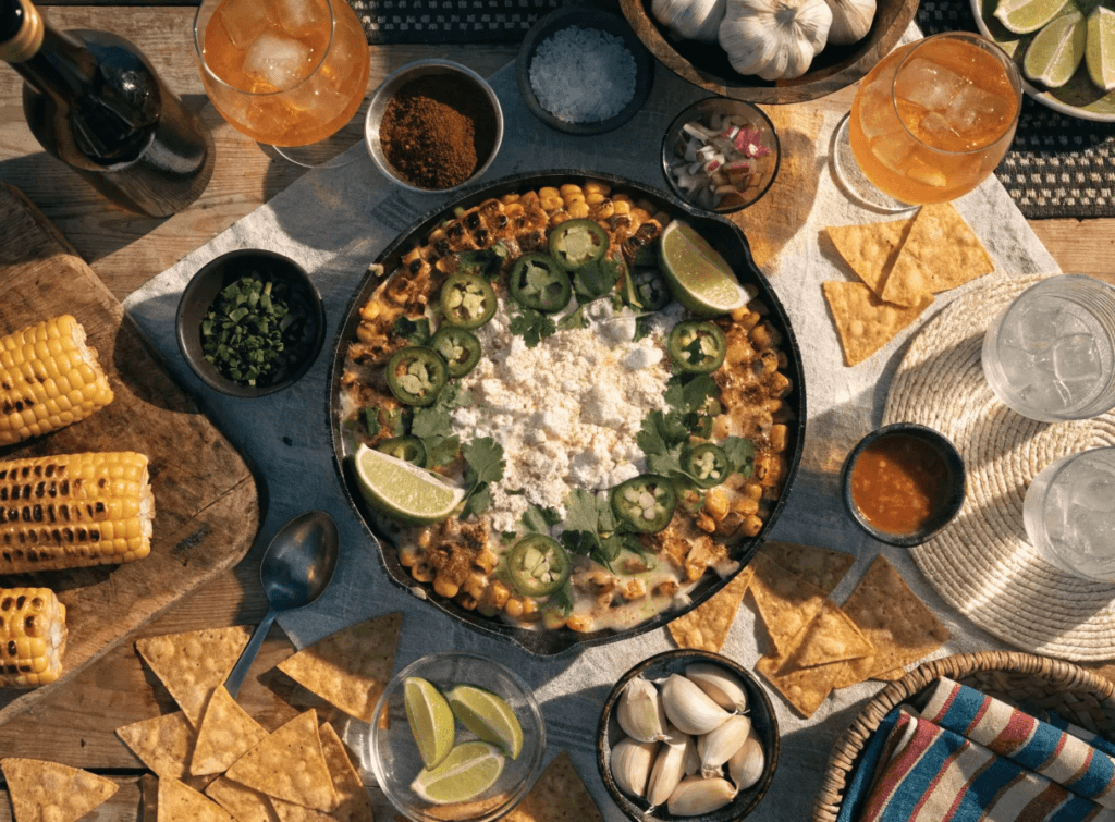 Zoomed-out presentation of fire-roasted elote dip surrounded by grilled corn, tortilla chips, lime wedges, garlic, drinks, and rustic BBQ table styling.