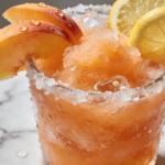 Frozen peach lemonade slush with frosty sugar rim, peach slices, lemon garnish, and crushed ice in a refreshing summer drink presentation.