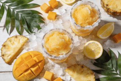 Top view of Frozen Mango Pineapple Lemonade slush drinks with mango cubes, pineapple slices, lemon wedges, and crushed ice on a tropical summer table.