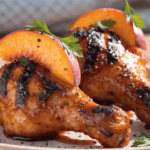 Close-up of grilled BBQ chicken drumsticks coated in sticky peach glaze with charred edges, fresh peach slices, parsley garnish, and glossy sweet smoky sauce.