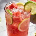 Close-up front view of Watermelon Lime Lemonade with fresh watermelon cubes, lime slices, lemon wedges, crushed ice, and chilled summer drink presentation.