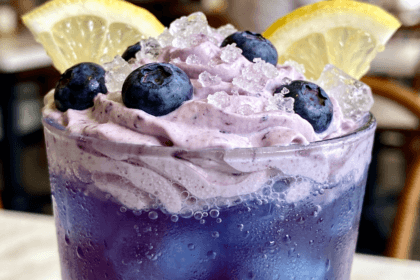 Close-up front view of creamy Whipped Blueberry Lemonade with fresh blueberries, lemon slices, whipped topping, and crushed ice in a frosted glass.