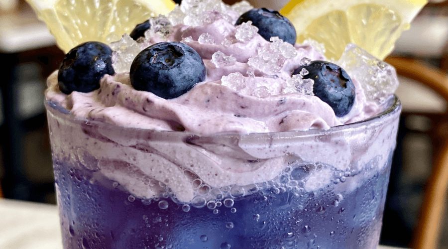 Close-up front view of creamy Whipped Blueberry Lemonade with fresh blueberries, lemon slices, whipped topping, and crushed ice in a frosted glass.