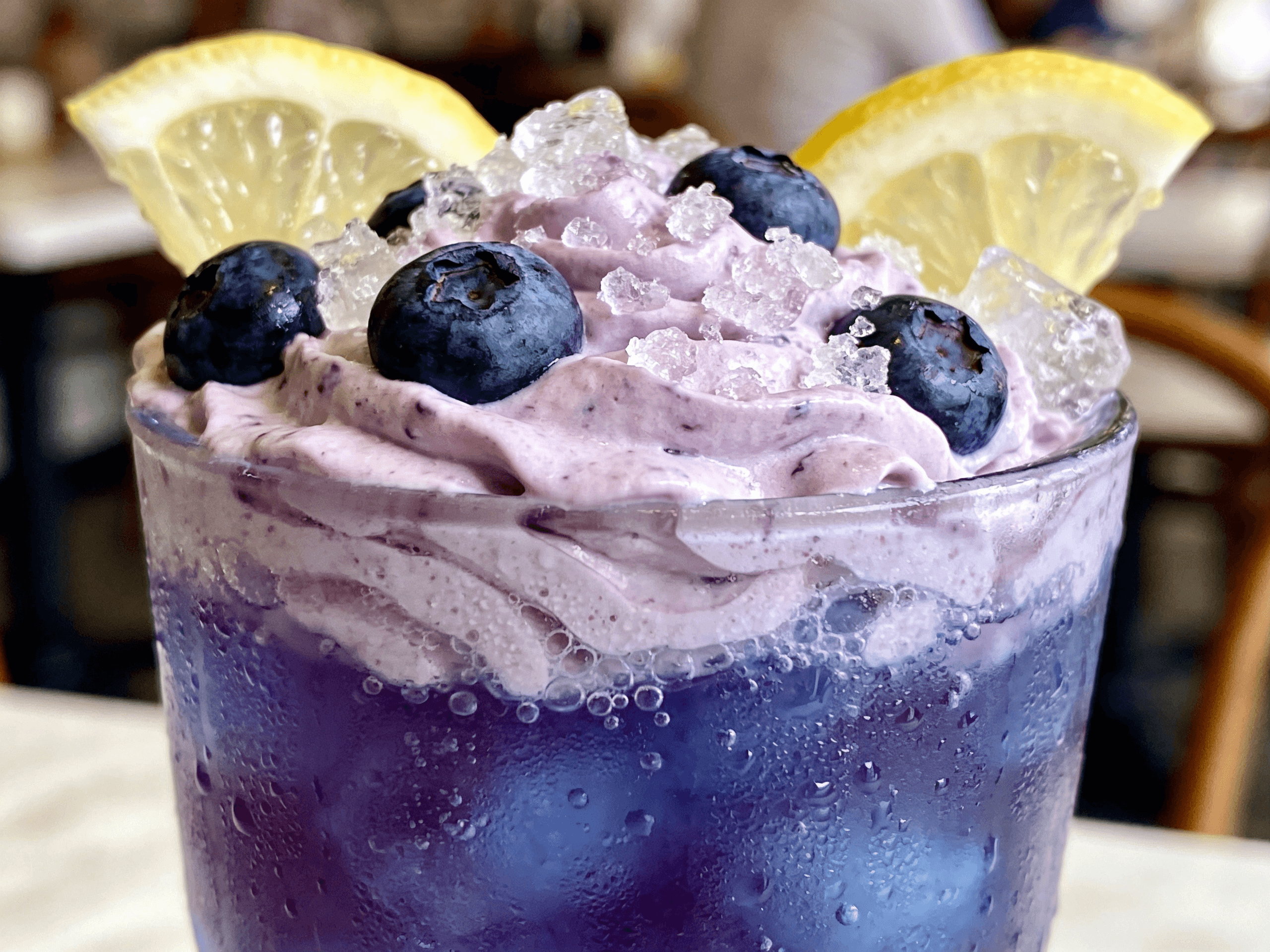 Close-up front view of creamy Whipped Blueberry Lemonade with fresh blueberries, lemon slices, whipped topping, and crushed ice in a frosted glass.