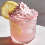Close-up front view of creamy Whipped Pink Lemonade with fluffy pink topping, fresh lemon slices, and ice in a frosted glass.