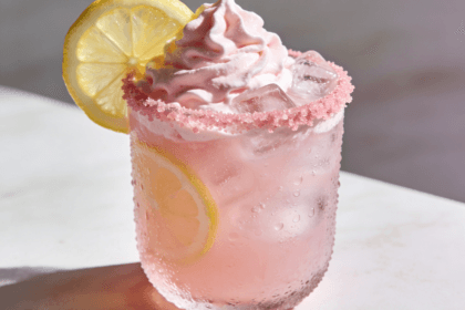 Close-up front view of creamy Whipped Pink Lemonade with fluffy pink topping, fresh lemon slices, and ice in a frosted glass.