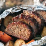 Close-up of grilled BBQ meatloaf foil packets with juicy meatloaf slices, roasted potatoes, carrots, and barbecue glaze.