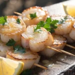 Close-up of grilled scallop skewers brushed with lemon garlic butter, parsley, and fresh lemon wedges.
