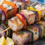 Close-up of Mediterranean grilled swordfish skewers with bell peppers, zucchini, red onion, lemon wedges, and fresh herbs.
